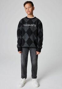 Ragazzo in piedi che indossa un sweater argyle nero e grigio con la scritta "ALLSAINTS", jeans scuri e sneakers bianche su uno sfondo grigio chiaro.