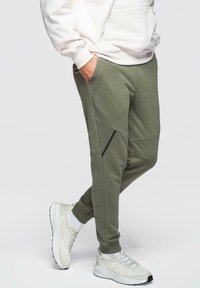 Person med hånden i lommen iført olivengrønne joggers og hvide sneakers, stående mod en ensfarvet baggrund.