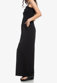 Kvinna som står sida vid sida iförd en ärmlös svart maxiklänning med smockad byst och händerna i sidofickorna, kompletterad med svarta plattformssandaler.