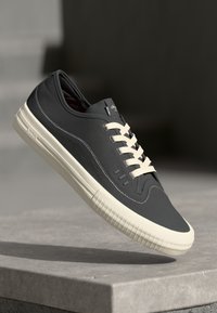 Mørkegrå sneakers med strukturert lærøvre, kremfargede snører og gummisåle med vertikale ribber. Minimalistisk design og sømdetaljer.