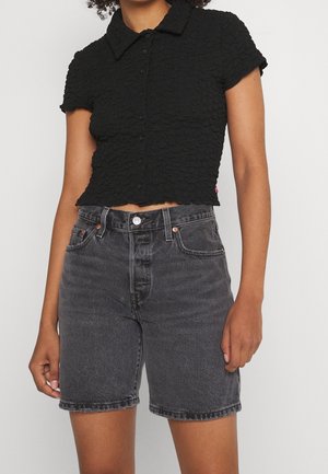 Woman wearing a black textured short-sleeve button-up cropped shirt and high-waisted faded black denim shorts.