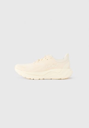 Zapatilla de running beige claro con parte superior de malla, collar acolchado, suela gruesa y amortiguada, y el logo de la marca en relieve en el lateral.