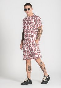 Muški komplet koji se sastoji od kratkih rukava, majice s crvenim geometrijskim uzorkom i kratkih hlača, izrađen od lagane tkanine. Crne tenisice upotpunjuju izgled.