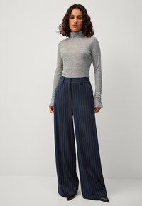 Grauer langärmliger Rollkragenpullover kombiniert mit hochgeschnittenen dunkelblauen Hose mit weißen Nadelstreifen und weiter Beinform.