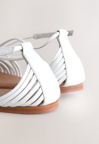 Sandales blanches à brides avec une semelle en bois plate. Les brides présentent un design tissé, créant un aspect texturé à l'arrière.