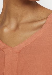 Haut à col en V en tissu pêche avec une texture subtile et une coupe décontractée, présentant un détail de couture nette le long de l'encolure et des épaules.