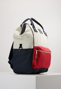 Plecak z kremowymi, granatowymi i czerwonymi sekcjami, z zewnętrzną kieszenią na zamek i skórzanym uchwytem; wykonany z wytrzymałego materiału z mocnymi uchwytami.