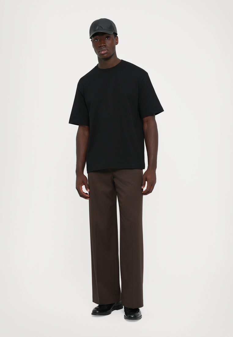 Homme portant une casquette noire, un t-shirt ample noir, un pantalon large marron et des chaussures noires, se tenant contre un fond blanc uni.