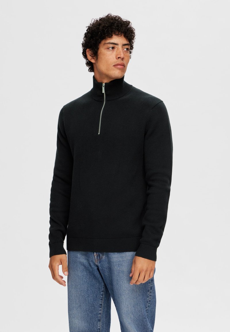 Black ribbed knit sweater with a high collar and half-zip closure; worn with light blue jeans, featuring long sleeves and a fitted design.