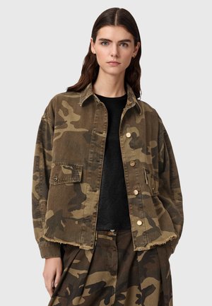 Młoda kobieta ubrana w oversize'ową zielono-brązową kurtkę moro i pasujące spodnie, pod którymi ma czarną koszulkę, stoi na tle gładkiego tła.