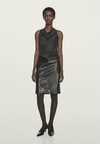 Haut en satin noir à col bénitier associé à une jupe crayon en cuir noir. Le modèle porte des collants noirs et des escarpins à bout pointu. Texture lisse, design épuré.