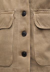 Μπεζ ύφασμα με τρεις μαύρα κουμπιά ευθυγραμμισμένα κατακόρυφα μεταξύ δύο θυλάκων με εμφανή ραφή.