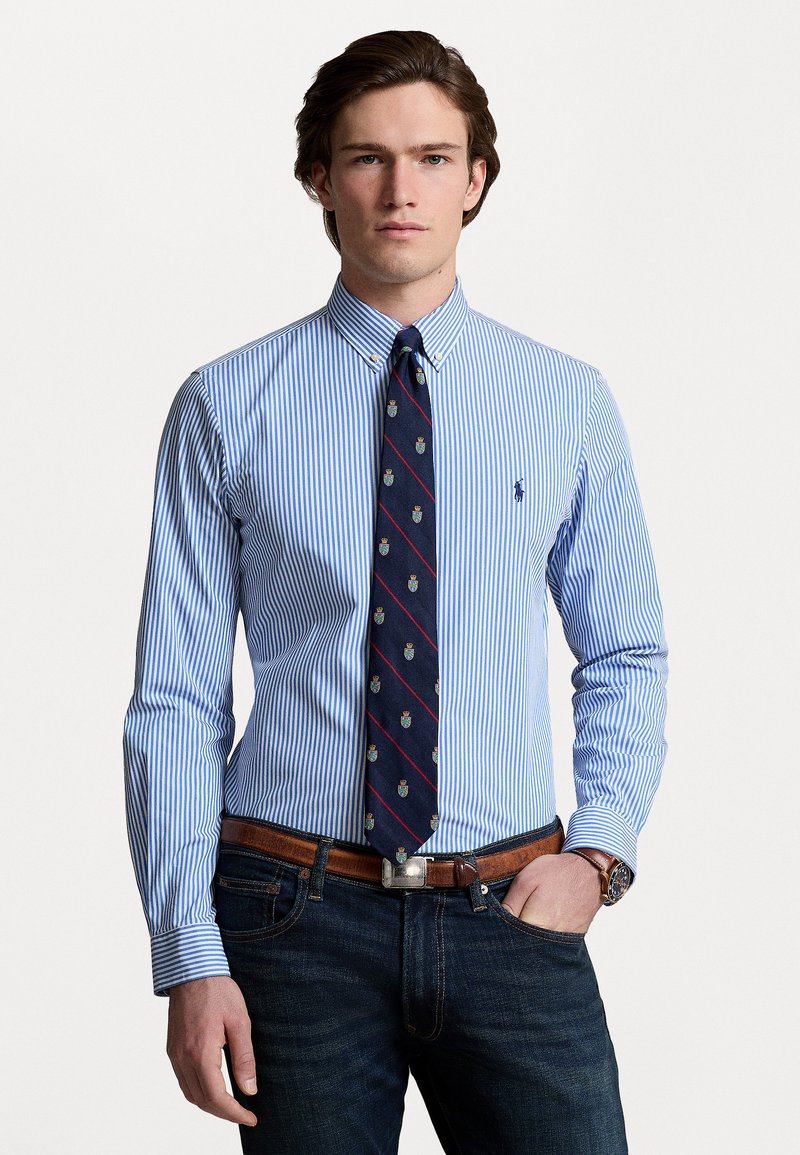 Camicia a righe blu e bianche con colletto, abbinata a una cravatta blu navy con motivi emblematici, jeans di denim e una cintura marrone.