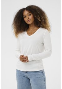 Weißes langärmliges V-Ausschnitt-Oberteil aus weichem Material, mit lässigem Schnitt und betonten Bündchen. Kombiniert mit hellblauen Denim-Jeans.