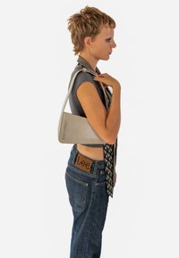 Borsa a spalla grigia, triangolare, con una superficie testurizzata e un marchio in rilievo. Indossata sopra una maglietta aderente e jeans blu con una toppa di marca.