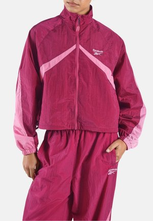 Person trägt eine winddichte Reebok-Jacke in Bordeauxrot und Pink sowie passende Hosen, hat eine Hand in der Tasche und zeigt einen neutralen Gesichtsausdruck.