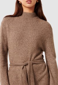 Brauner, gestrickter Pullover mit hohem Kragen. Verfügt über einen Bindegürtel in der Taille und lange Ärmel, hergestellt aus einem weichen, strukturierten Material.
