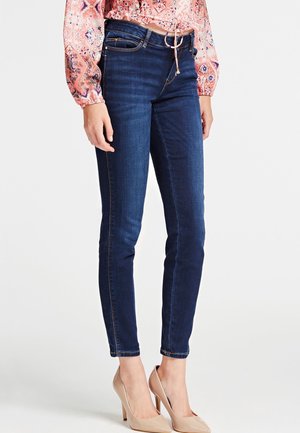 Woman wearing dark blue skinny jeans, nude high heels, and a pink patterned long-sleeve blouse, standing against a plain white background.