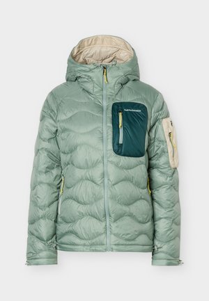 Veste matelassée vert clair avec capuche, poche poitrine vert foncé, poche sur la manche beige, tirettes de fermeture éclair jaunes, et fermeture éclair frontale.