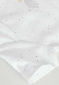 Weißes Baumwollkleidungsstück mit kleinen grauen Pünktchen, abgerundetem Saum und weicher Textur. Sichtbar bedruckte Grafik in der Nähe der Schulter.