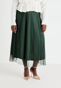 Gonna maxi plissettata verde con un sottovestito trasparente, abbinata a scarpe con tacco nude con un accento metallico. Blusa bianca con volant.