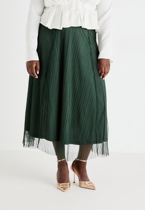 Jupe maxi plissée verte avec un overlay transparent, associée à des chaussures à talons nus avec un accent métallique. Blouse blanche avec des volants.