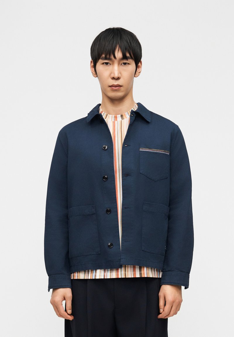 Νέος άνδρας φοράει μπλε σκούρο σακάκι με κουμπιά πάνω από πολύχρωμο ριγέ πουκάμισο, στέκεται μπροστά σε απλό λευκό φόντο.