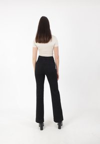 Jeans negros acampanados de cintura alta, combinados con una blusa ajustada de color beige claro. La modelo está de espaldas frente a la cámara sobre un fondo blanco liso.