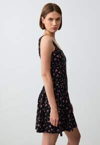 Zwarte bloemenjurk met dunne bandjes, met een aansluitend bovenstuk en een uitlopende rok. Klein patroon van roze bloemen en groene bladeren over de hele jurk.
