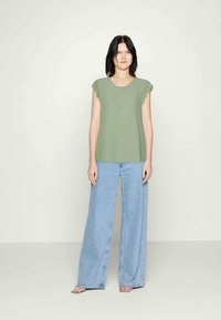 Blusa verde de mangas curtas com bordas franzidas, combinada com calças de ganga largas na cor azul clara e sapatos com padrão preto e branco. Fundo simples.