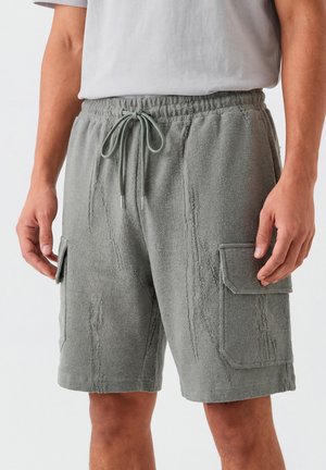 Hombre vistiendo pantalones cortos cargo grises con textura, cintura con cordón y bolsillos con solapa, combinados con una camiseta blanca.