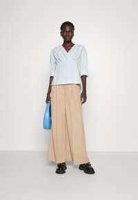 Lys blå, stripete bluse med dyp V-hals og rynket midje, parret med beige vide bukser og svarte platåsko. Holder en blå veske.
