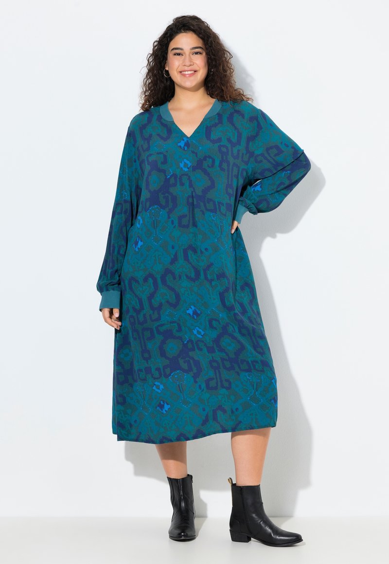 Vestido largo y holgado en tonos azul verdoso y azul oscuro con patrones geométricos. Presenta un escote en V y mangas largas. Se lleva con botas negras hasta el tobillo.