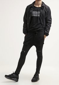 Svart outfit bestående av en zip-up jacka, en grafisk t-shirt med "BLACK KAT" och åtsittande byxor med blixtlås. Avslutad med svarta sneakers.