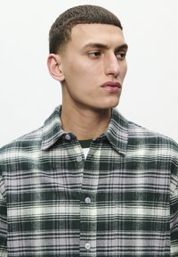 Green and white plaid flannel shirt with a button-up design, featuring a collar, chest pocket, and subtle logo detail. Textured fabric.