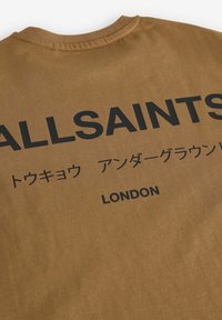 Brūna kokvilnas krekls ar izteiksmīgu melnu tekstu, kas vēsta "ALLSAINTS" un "LONDON", ar papildus tekstu japāņu rakstzīmēs.