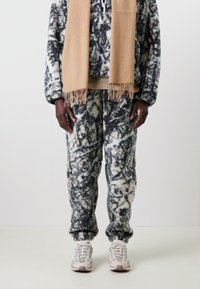 Fleece hoodie and pants in black and white abstract print, paired with a beige fringed scarf. White sneakers complete the outfit.