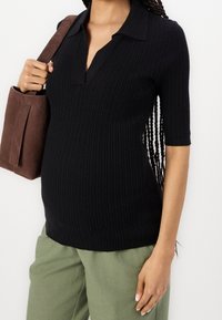 Czarna żakardowa bluzka z kołnierzykiem i krótkimi rękawami, zestawiona z oliwkowymi spodniami. Brązowa zamszowa torba trzymana pod pachą.