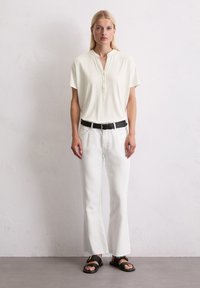 Blouse à manches courtes crème avec col et patte de boutonnage, associée à un jean blanc évasé et des sandales noires. Design simple et moderne.