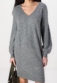 Robe tricotée grise avec décolleté en V, manches longues bouffantes et ourlet côtelé ; texture lisse et coupe décontractée. Accompagnée d'un sac à épaule vert.