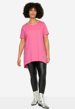 Rosa Baumwolltunika mit kurzen Ärmeln, Rundhalsausschnitt und asymmetrischem Saum, kombiniert mit schwarzen Leggings und silbernen Reißverschluss-Stiefeletten.