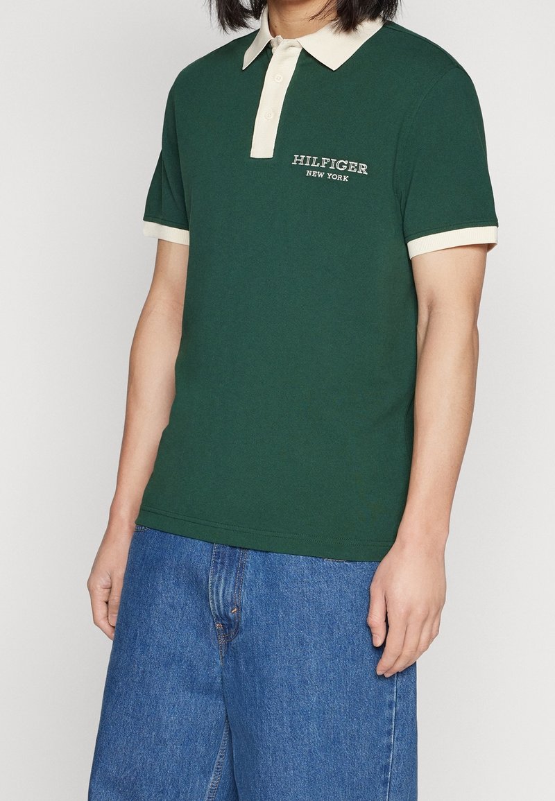 Personne portant un polo vert foncé avec un col et des poignets beiges, logo "Hilfiger New York" sur la poitrine, associé à un jean bleu décontracté.