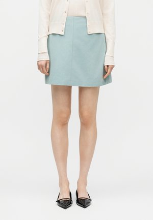 Vrouw die een lichtblauwe suède minirok draagt, een beige vest met knopen en zwarte pumps met een puntige neus, staand tegen een witte achtergrond.