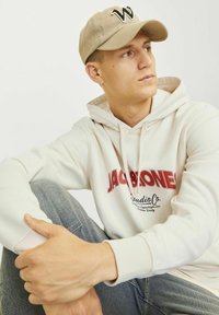 Gorra de béisbol beige con un oscuro logo "W", sudadera crema con logo y texto en rojo, jeans gris claro. Pose relajada sentado, con una mano en la rodilla.