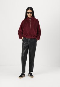 Burgunderfarget fleece-pull med høy krage og kvart-glidelås, kombinert med svarte imitert lærbukser og svarte sneakers med hvite striper.