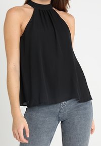 Svart halterneck-topp i lätt tyg, med hög halsringning, lös passform och utsvängd nedre kant, ihop med blå jeans.