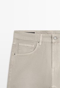 Pantalon beige avec poche avant et poche pour pièce de monnaie, passants pour ceinture, fermeture par bouton métallique, et détails de couture apparents.