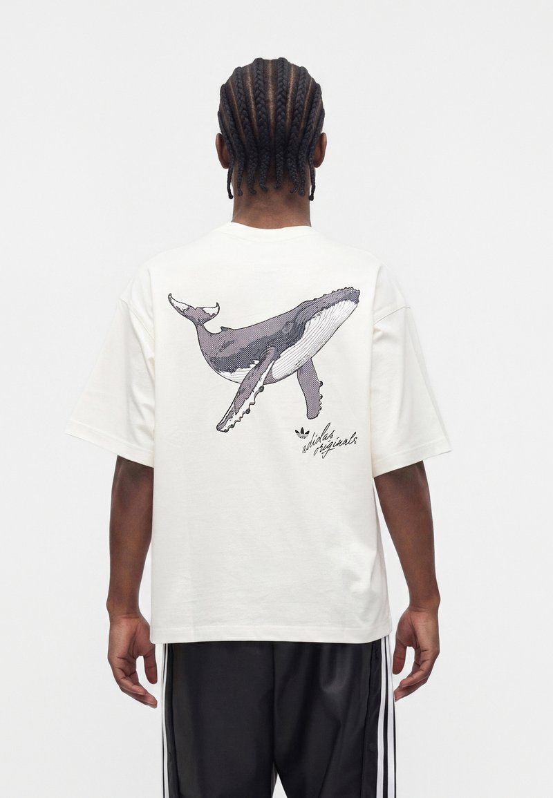 Homme aux cheveux tressés portant un t-shirt blanc avec un motif graphique de baleine en noir et blanc et le texte « adidas originals » dans le dos.