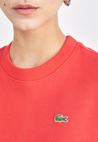 Sarkans kokvilnas svārsts ar apaļu kakla izgriezumu un teksturētu apdari. Iezīmēts ar mazu zaļu krokodilu logotipu kreisajā krūtī.