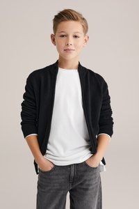 Svart stickad cardigan med dragkedja och ribbade muddar över en vit t-shirt, kombinerad med mörkgrå jeans. Slät textur och avslappnad passform.